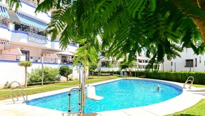 Piscina con trampolín y silla para salvavidas, rodeada de un césped cuidado y un edificio con balcones.