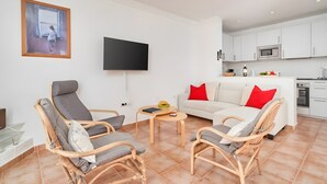 Un salon avec des fauteuils en osier, un canapé blanc agrémenté de coussins rouges, un téléviseur à écran plat et un coin cuisine avec des meubles et des appareils électroménagers blancs.