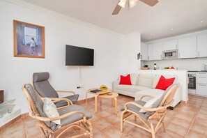 Un salon avec des fauteuils en osier, un canapé blanc agrémenté de coussins rouges, un téléviseur à écran plat et un coin cuisine avec des meubles et des appareils électroménagers blancs.