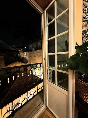 Vista nocturna desde un balcón con barandilla de metal, una ventana y una planta en maceta.