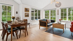 Une pièce spacieuse comprenant une table à manger en bois, des chaises, un canapé et une table basse. De grandes fenêtres laissent entrer la lumière naturelle et offrent une vue sur la verdure environnante.