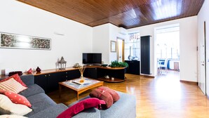 Un salón moderno con un sofá gris, suelo de madera, un televisor y una zona de comedor visible a través de una puerta abierta.