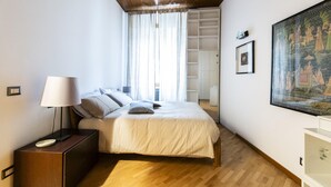 Un dormitorio con techo de madera, una cama con ropa de cama blanca, una mesita de noche con lámpara, un cuadro grande en la pared y un armario con estantes.