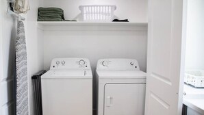 Uma lavanderia com duas máquinas de lavar brancas, um cesto na prateleira e uma porta que dá para outro cômodo.