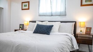 Uma cama bem arrumada com roupa de cama branca e um travesseiro azul, com mesas de cabeceira laterais com luminárias e quadros emoldurados.