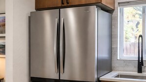 Un refrigerador de acero inoxidable con dos puertas y un compartimento congelador, ubicado en una cocina con gabinetes de madera y un ventanal.