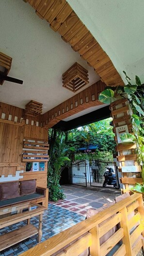 Un interior de madera con un banco y una mesa, un ventilador de techo y vistas a un jardín a través de un arco abierto.