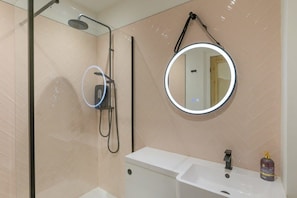 Un baño con ducha de mampara de vidrio, un espejo redondo con luz y un lavabo blanco.