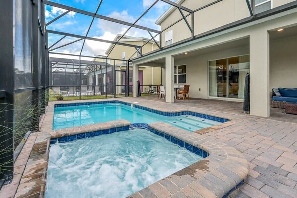 A swimming pool with a hot tub, surrounded by a covered patio area with seating and a glass enclosure.