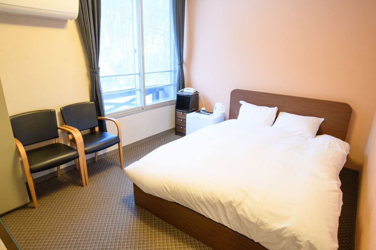 A hotel room with a bed, a chair, a small table with a lamp, and a window with curtains.
