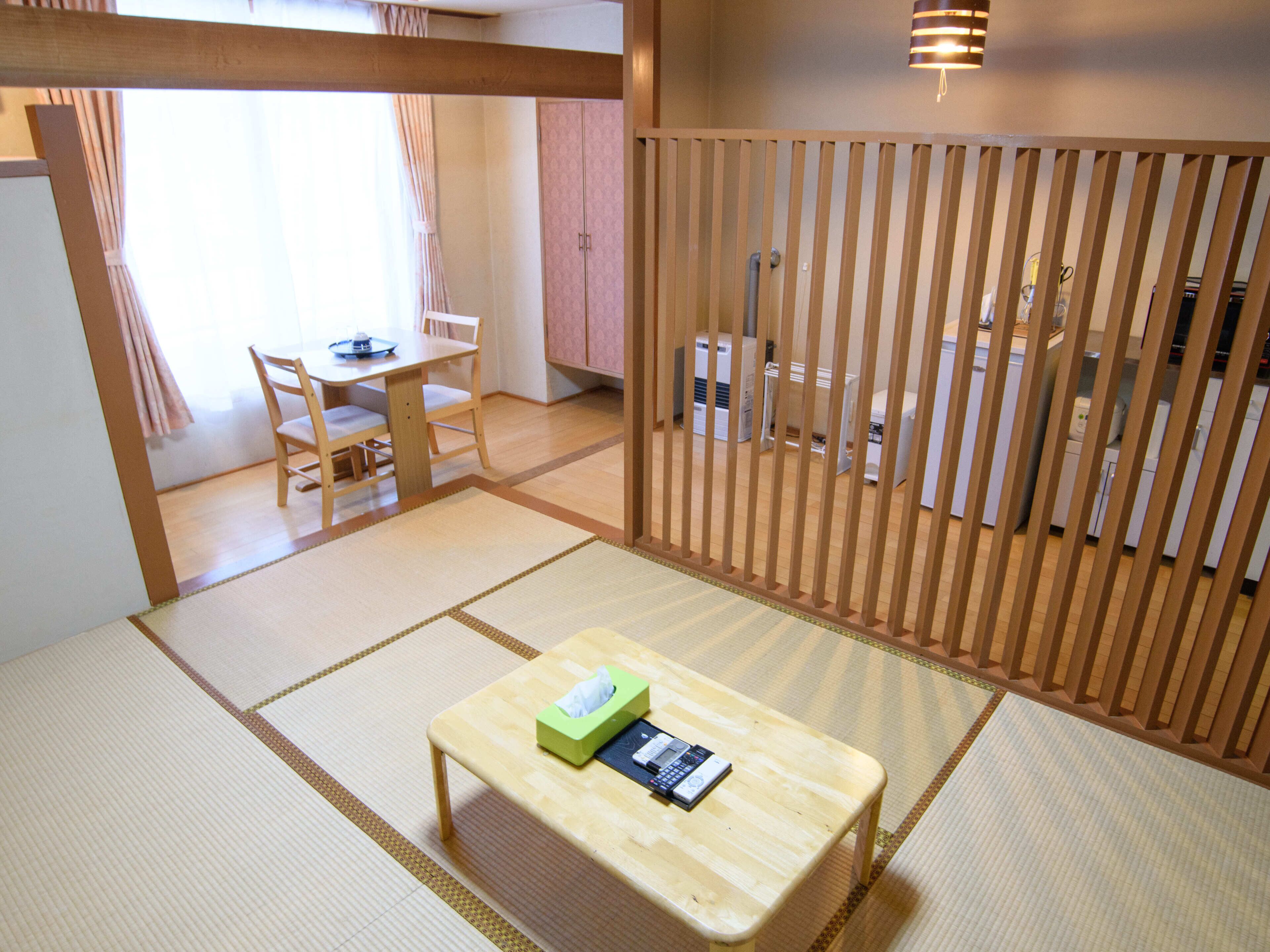 A traditional Japanese-style room with tatami flooring, a low wooden table with a tissue box and remote control, a wooden partition, and a view of a kitchen area.