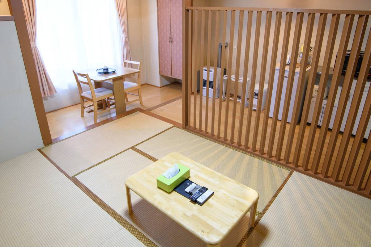 A traditional Japanese-style room with tatami flooring, a low wooden table with a tissue box and remote control, a wooden partition, and a view of a kitchen area.