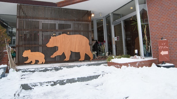 A building entrance with a wooden bear cutout, a brick facade, and a glass door. - ryobou hanaemi (Otaru)