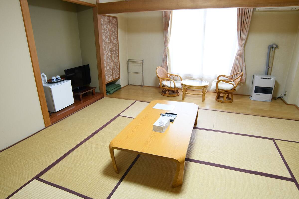 A room with tatami flooring, a wooden table, two wicker chairs, a television, a small cabinet, and a heater.