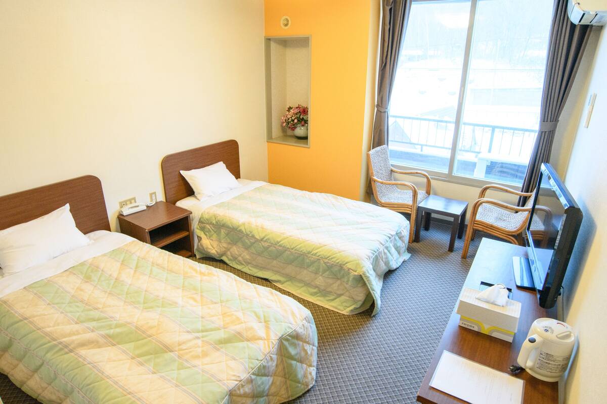 A hotel room with two beds, a small table, a chair, a desk, and a window with curtains.
