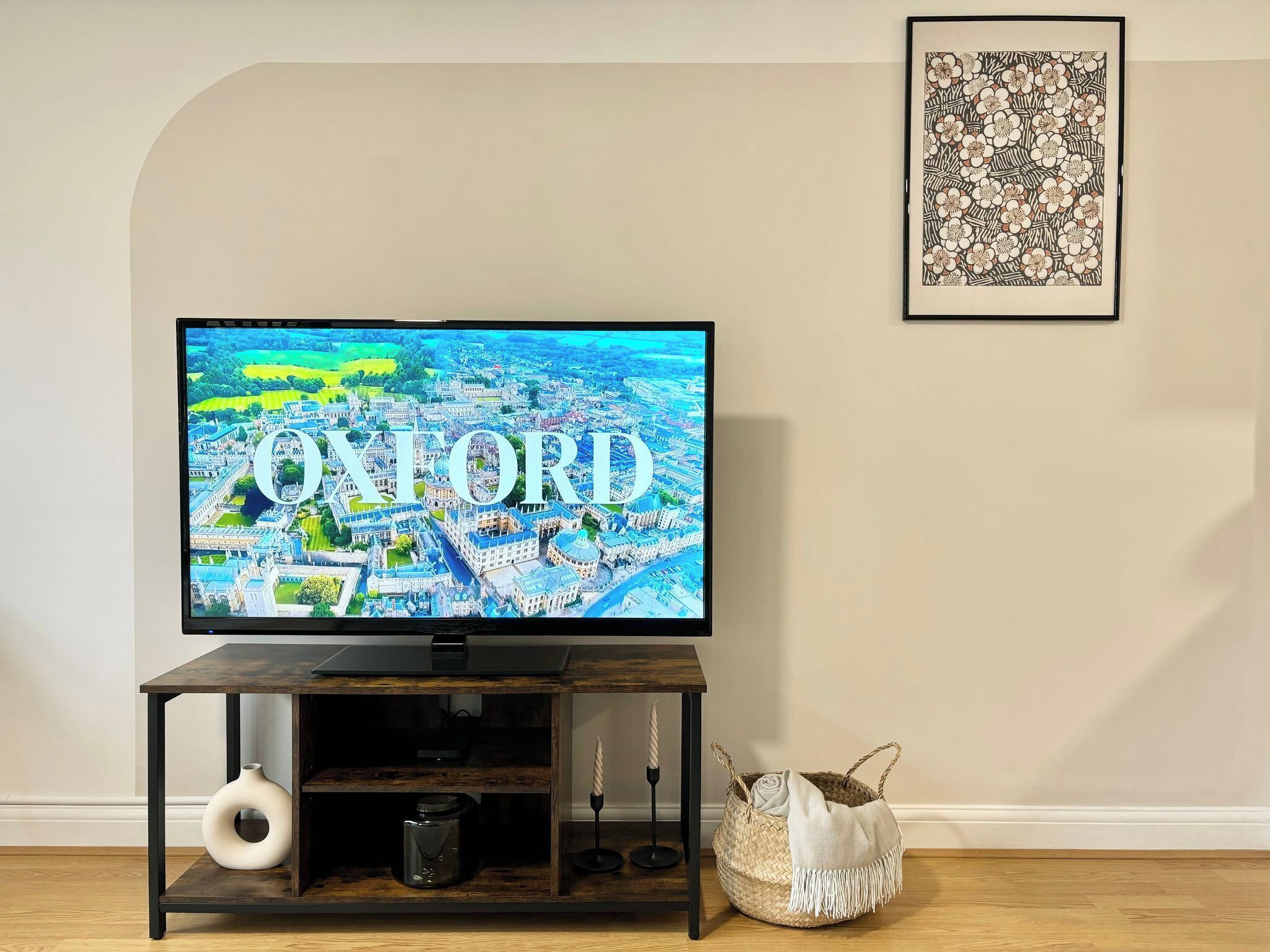 A television displaying an aerial view of Oxford, a framed artwork, a woven basket, and a candle holder on a wooden stand.