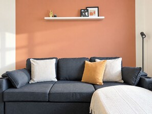 En moderne stue med en mørkegrå sofa, hvide og sennepsgule puder, et hvidt plaide og en minimalistisk hylde med pynteting.