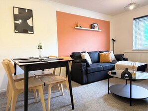 En moderne stue med en sort sofa, et spisebord med stole, et lille sofabord og en vægmonteret reol med pynteting.