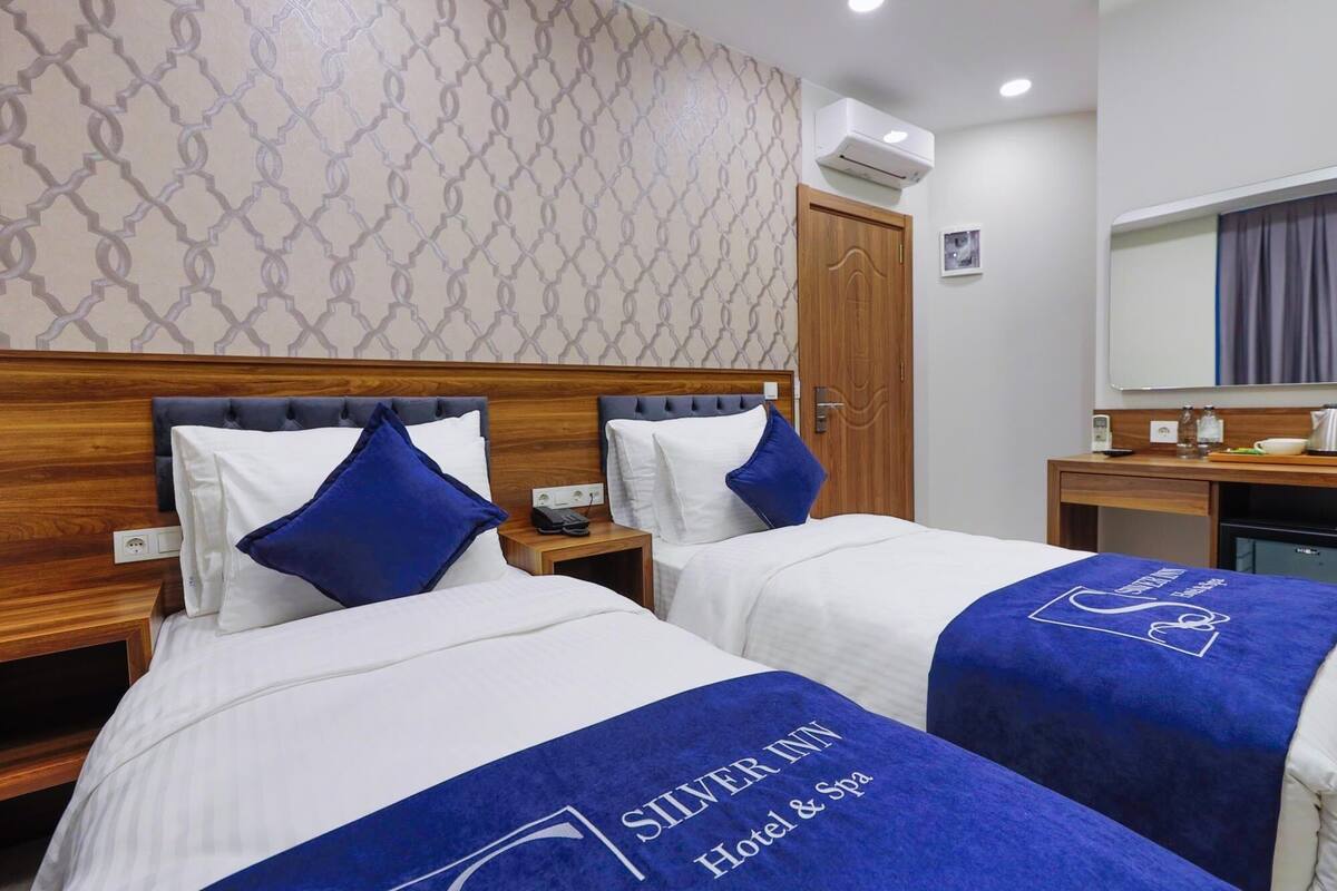 Une chambre d’hôtel avec deux lits, chacun recouvert d’une courtepointe bleue et doté d’oreillers, une tête de lit en bois, une table de chevet avec un téléphone et une lampe, et un miroir au-dessus de la table.