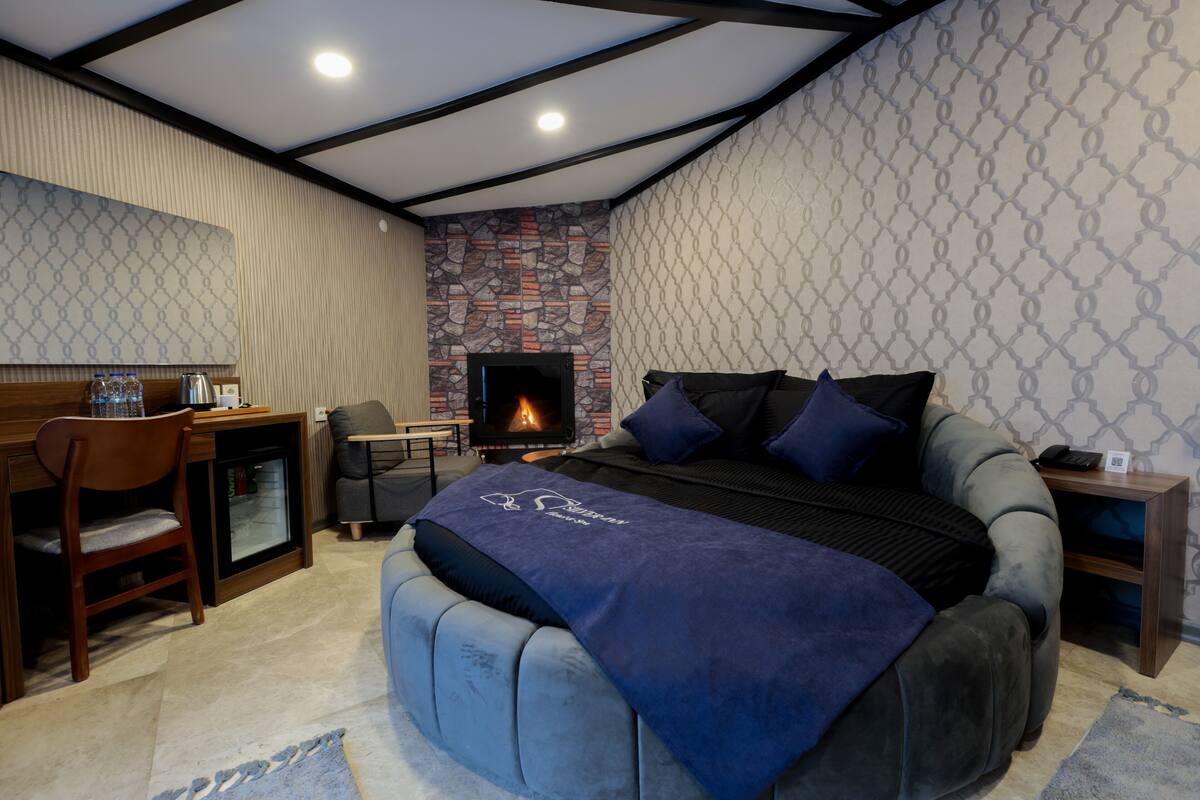 Une chambre avec une cheminée en pierre, un lit recouvert d’une couverture bleue, un bureau avec une chaise et une petite table avec un téléphone.