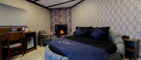 A bedroom with a stone fireplace, a bed with a blue blanket, a desk with a chair, and a small table with a phone.