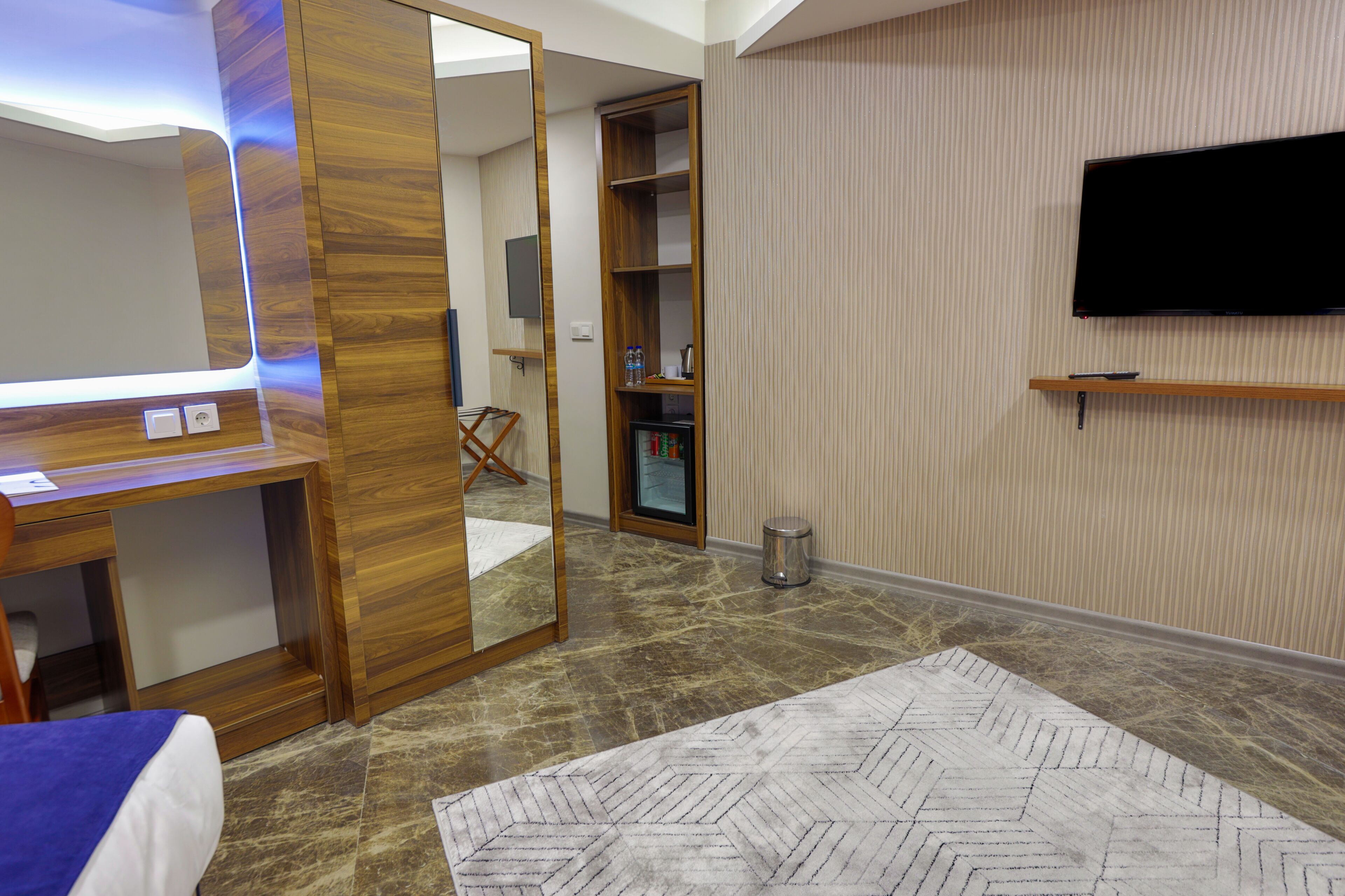 A hotel room with a wooden desk, a large mirror, a television mounted on the wall, a storage cabinet, and a patterned rug.
