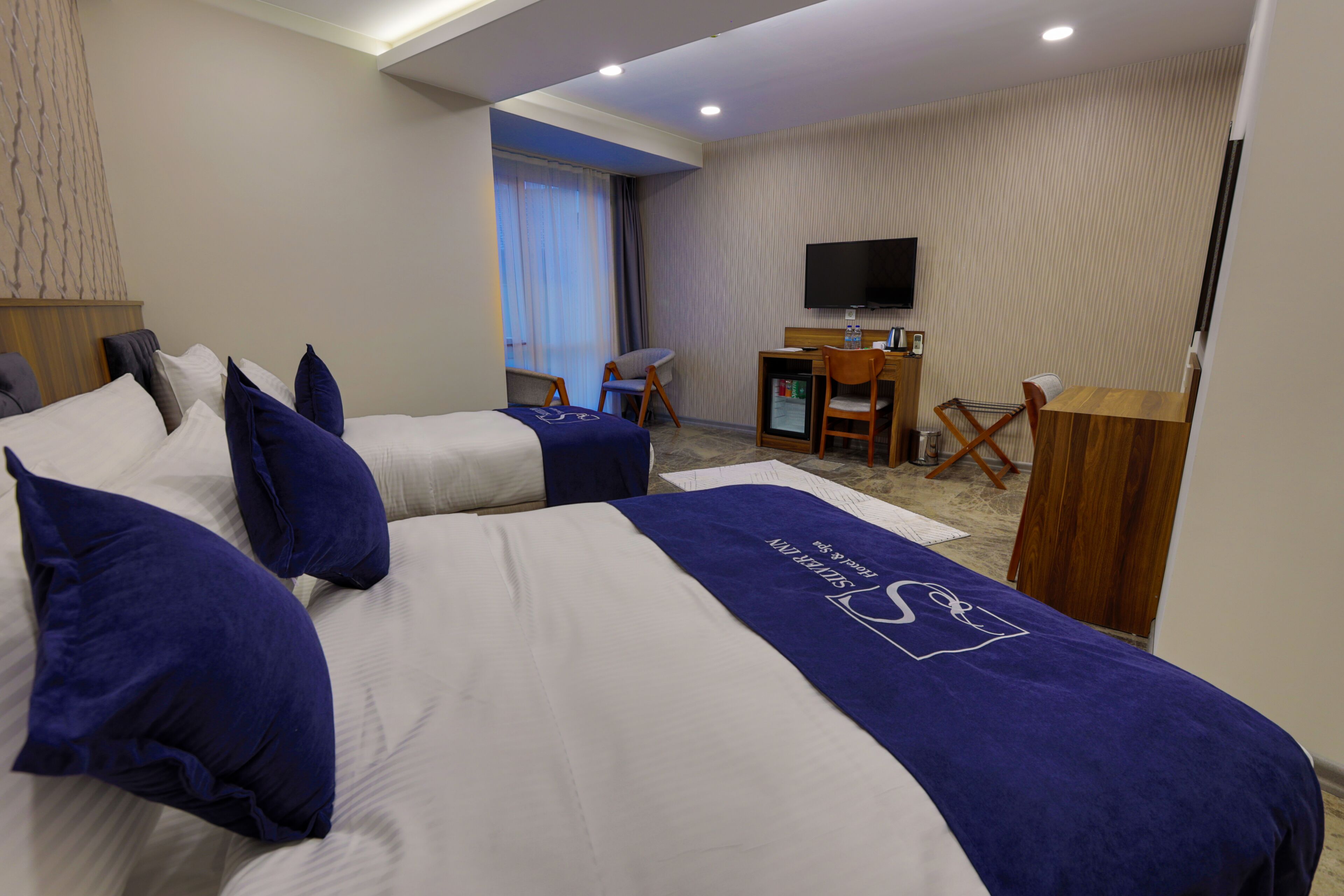 A hotel room with two beds, a desk, a chair, a television, and a minibar.