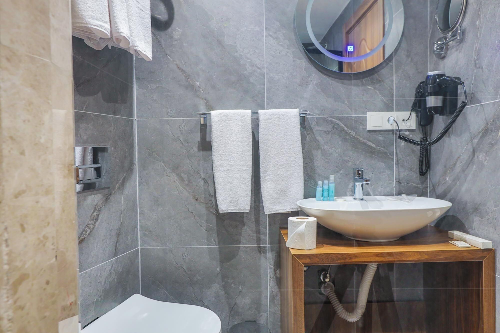 Un baño moderno con pared de mármol, un espejo redondo, tocador de madera y un inodoro.