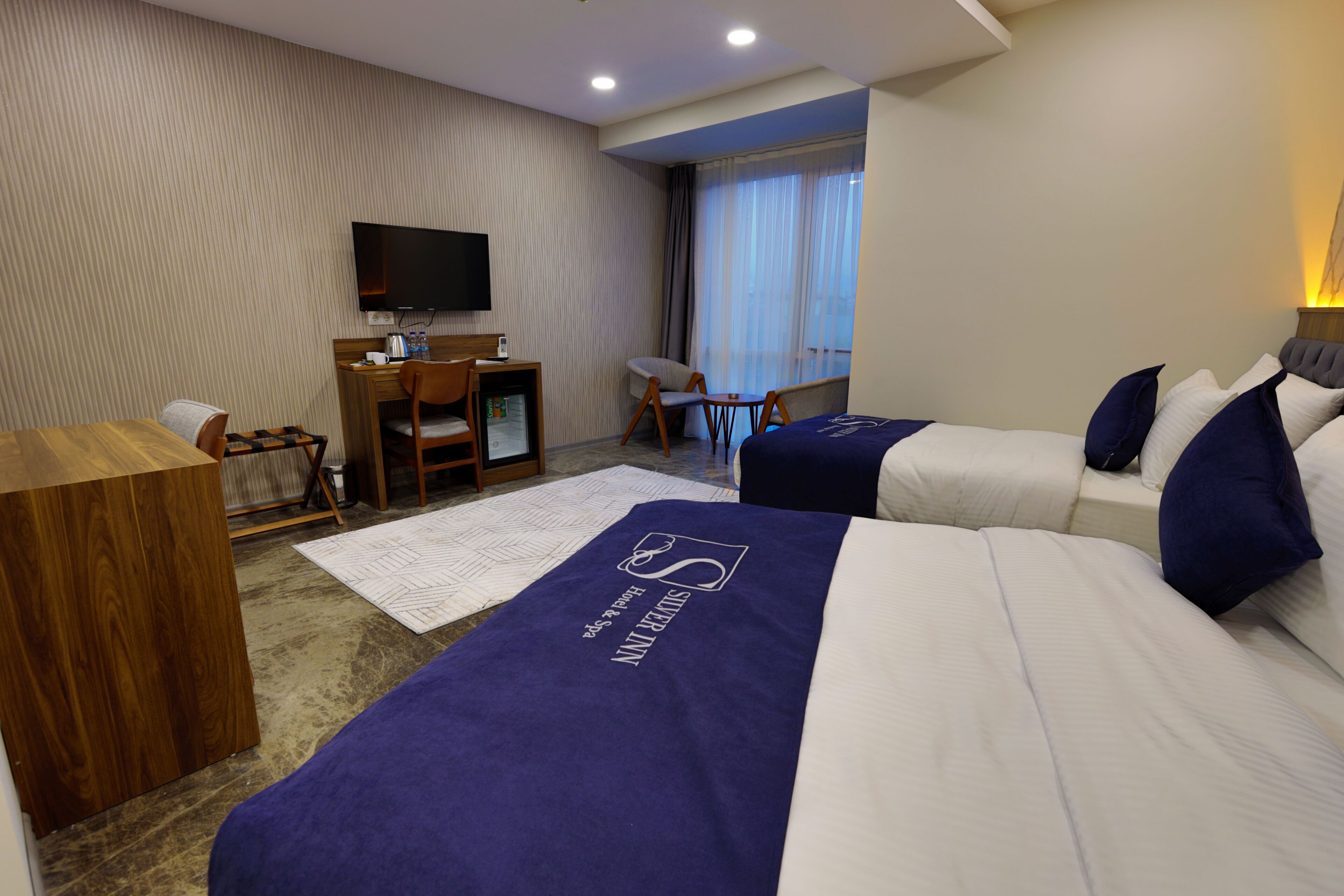 Quarto de hotel com duas camas, uma escrivaninha, uma cadeira, televisão e janela com cortinas.