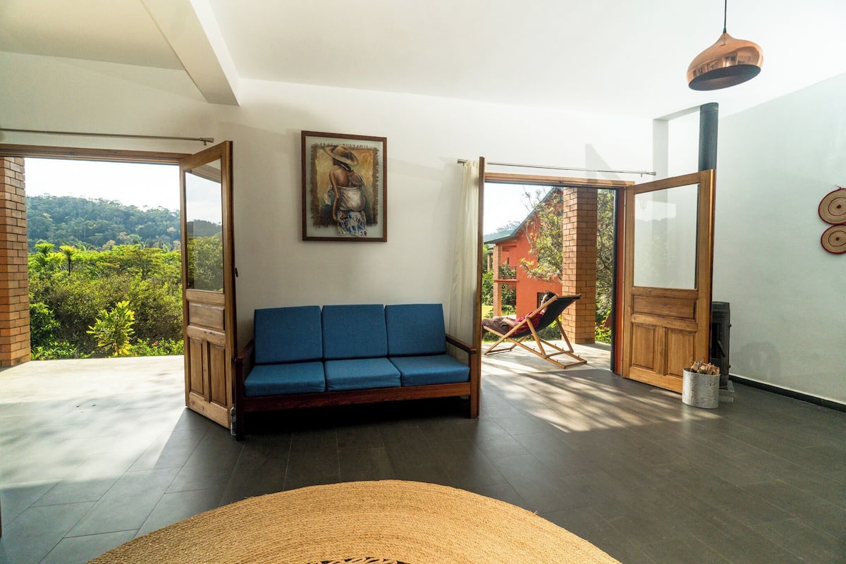 Uma sala de estar moderna com um sofá azul, um quadro na parede e vista para uma paisagem verde exuberante através de portas abertas.