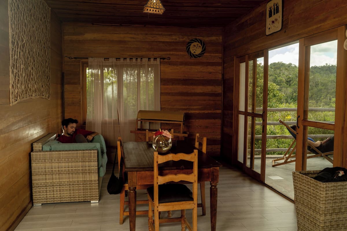 Interior de cabana de madeira com área de jantar, sofá de vime e vista para a floresta por grandes janelas.