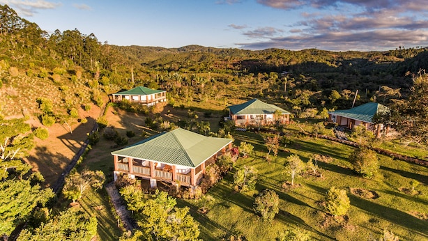 A large estate with three main buildings, each with a green roof, surrounded by lush greenery and hills in the background. - Eulophiella (Andasibe)