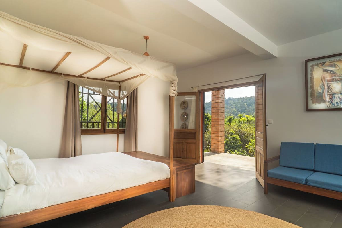 Quarto com cama, um sofá azul e vista para o verde pela janela.