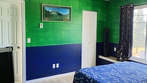 A room with green and blue walls, a bed with a blue patterned bedspread, a framed picture on the wall, and a window with curtains.