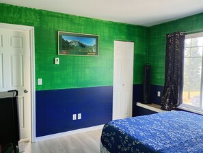 A room with green and blue walls, a bed with a blue patterned bedspread, a framed picture on the wall, and a window with curtains.