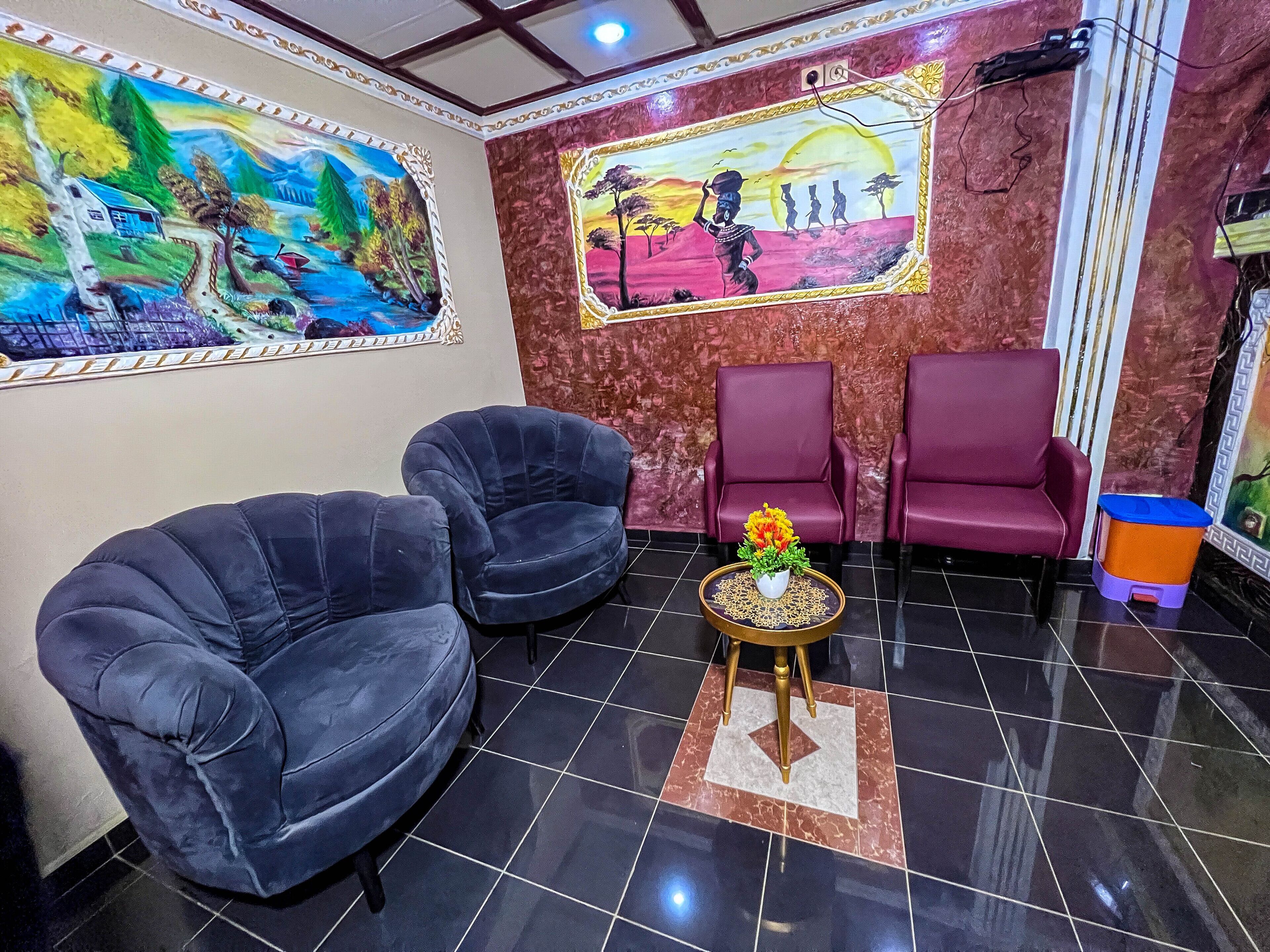 A reception area with two black armchairs, a small round table with a flower vase, and two red armchairs. There are two large paintings on the walls depicting landscapes and a sunset scene.