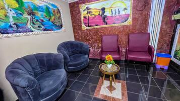 A reception area with two black armchairs, a small round table with a flower vase, and two red armchairs. There are two large paintings on the walls depicting landscapes and a sunset scene.