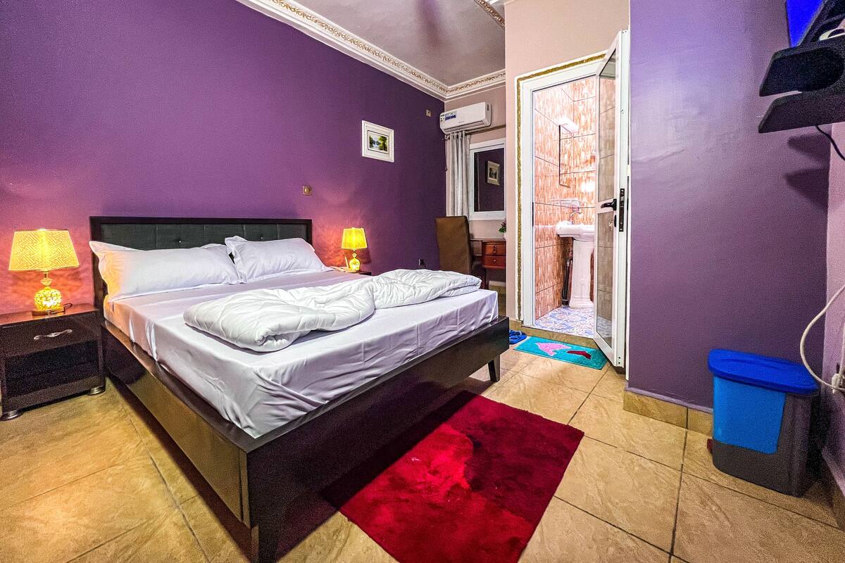 Un dormitorio con una cama, dos lámparas de noche, un tapete rojo, un cubo de basura azul y una puerta de baño.