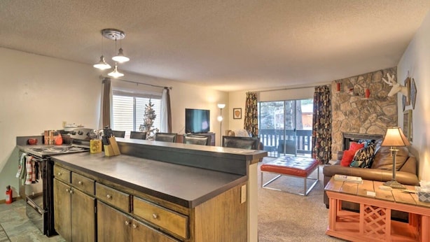 A spacious living area with a large kitchen island, a fireplace, and a balcony with a stone wall. - Angel Fire Condo Mountain Retreat by Velo (Angel Fire)