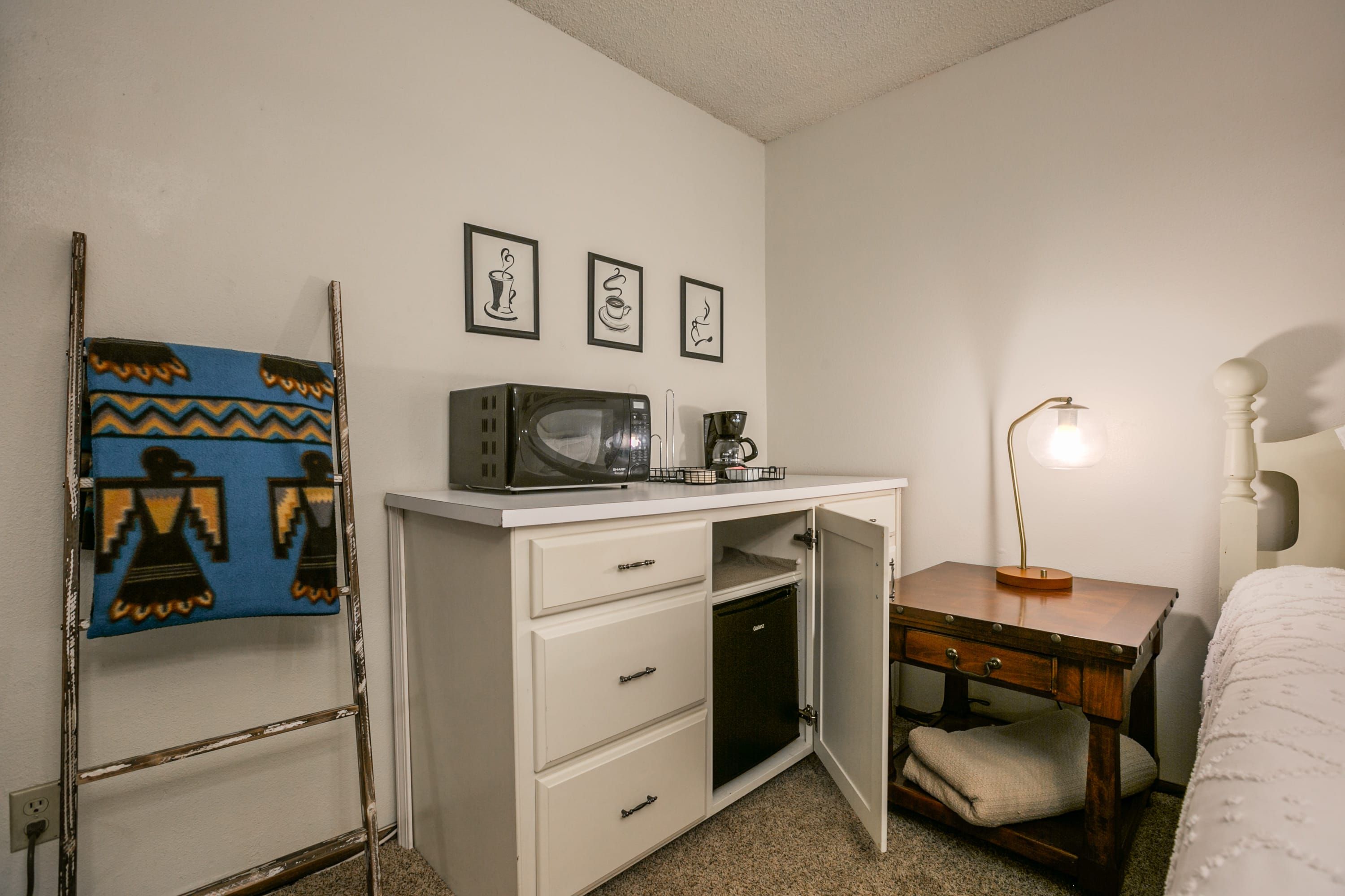 A compact bedroom with a bed, a desk with a computer and a coffee maker, a lamp, and a wall with three framed pictures.