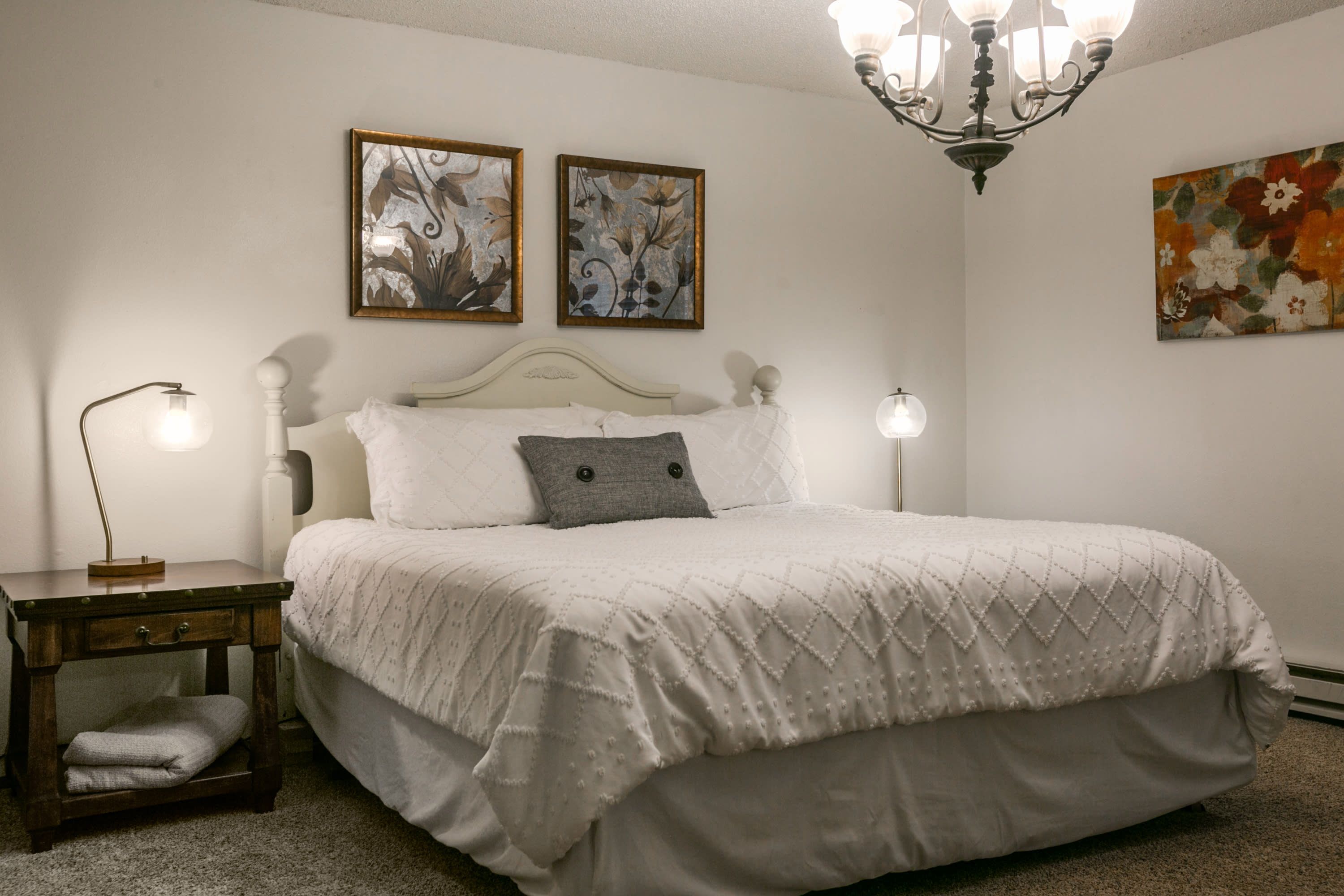 Quarto com cama, duas pinturas penduradas na parede, mesa de cabeceira com luminária e lustre.