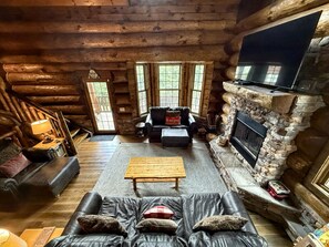 Un acogedor interior de cabaña de troncos con chimenea de piedra, un televisor de pantalla plana grande, sofás de cuero y una mesa de centro de madera.