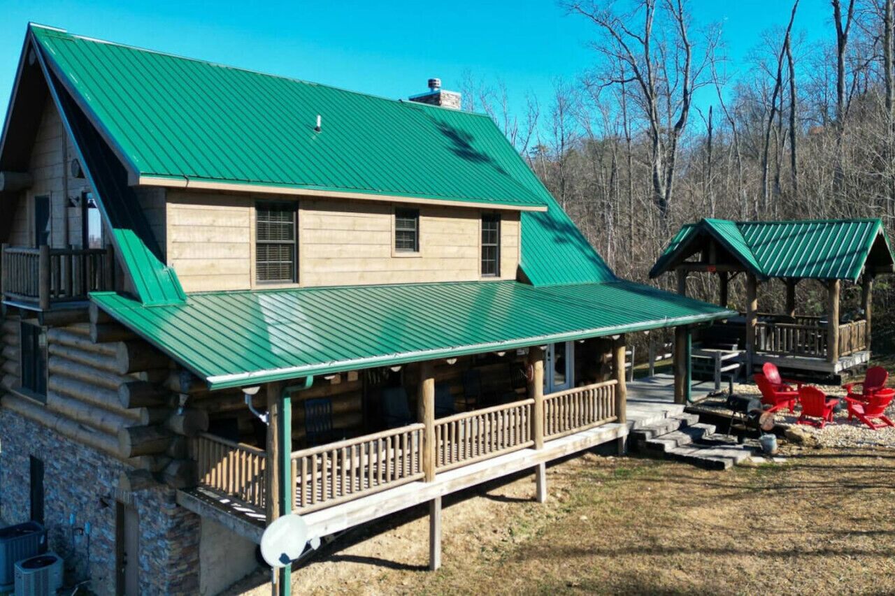 Une cabane en rondins surmontée d’un toit en métal vert, dotée d’une véranda avec des balustrades et d’une petite structure en bois également surmontée d’un toit vert.