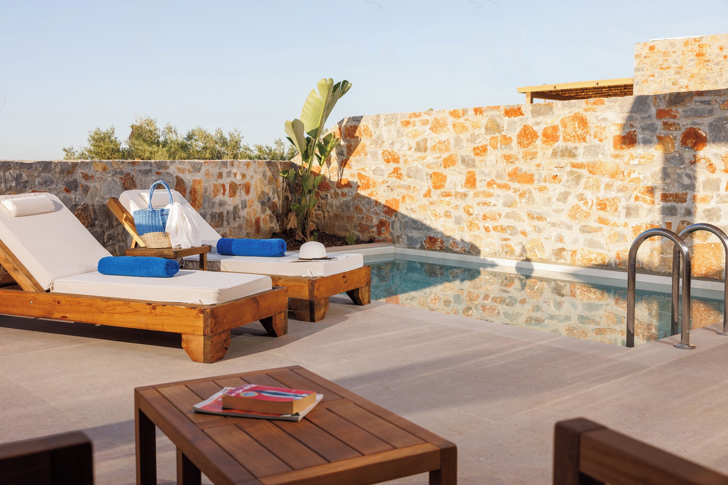 A poolside area with wooden loungers, a small table, and a stone wall.