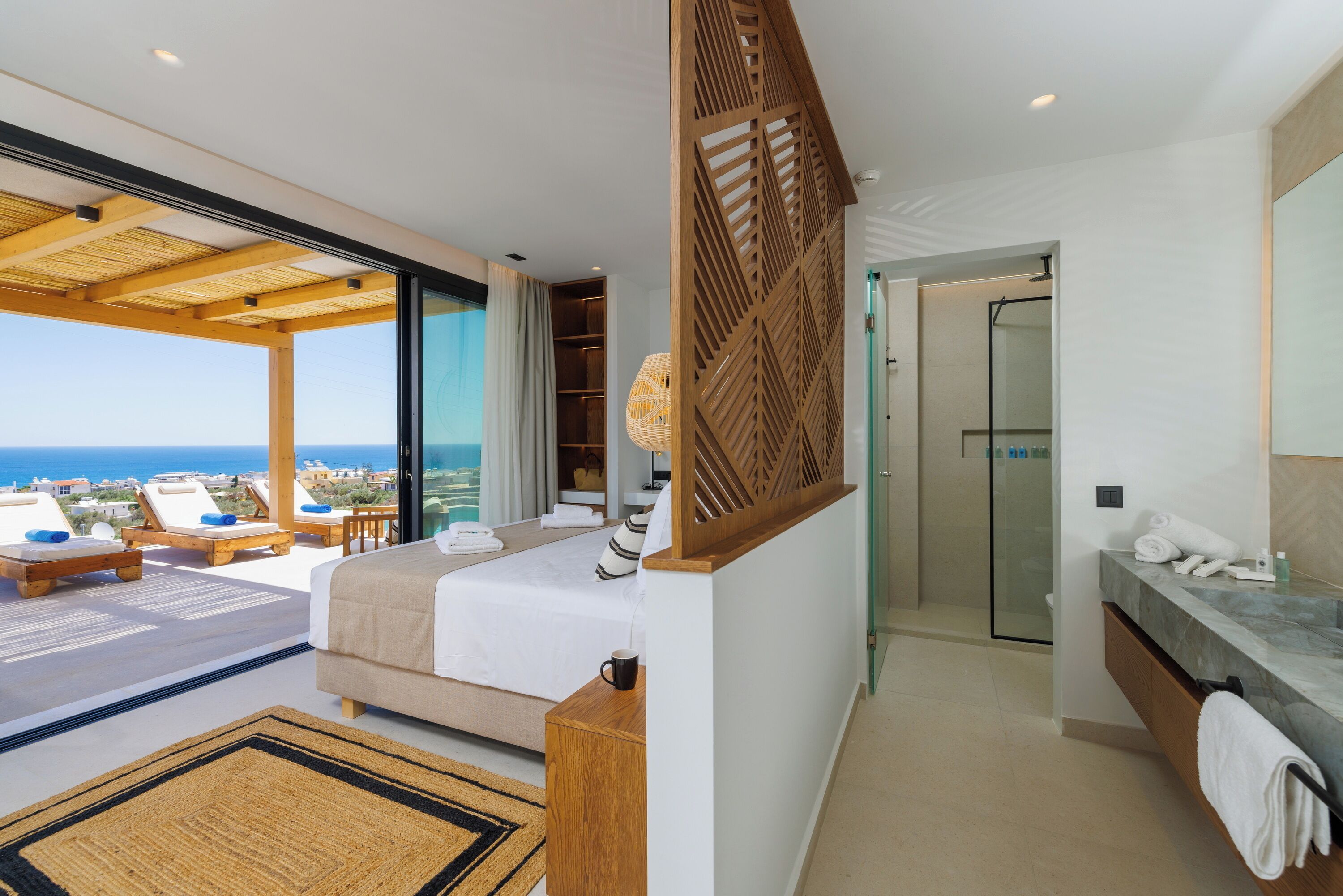 A modern bedroom with a large bed, a wooden partition, a bathroom with a glass shower, and a balcony with lounge chairs and a view of the sea.