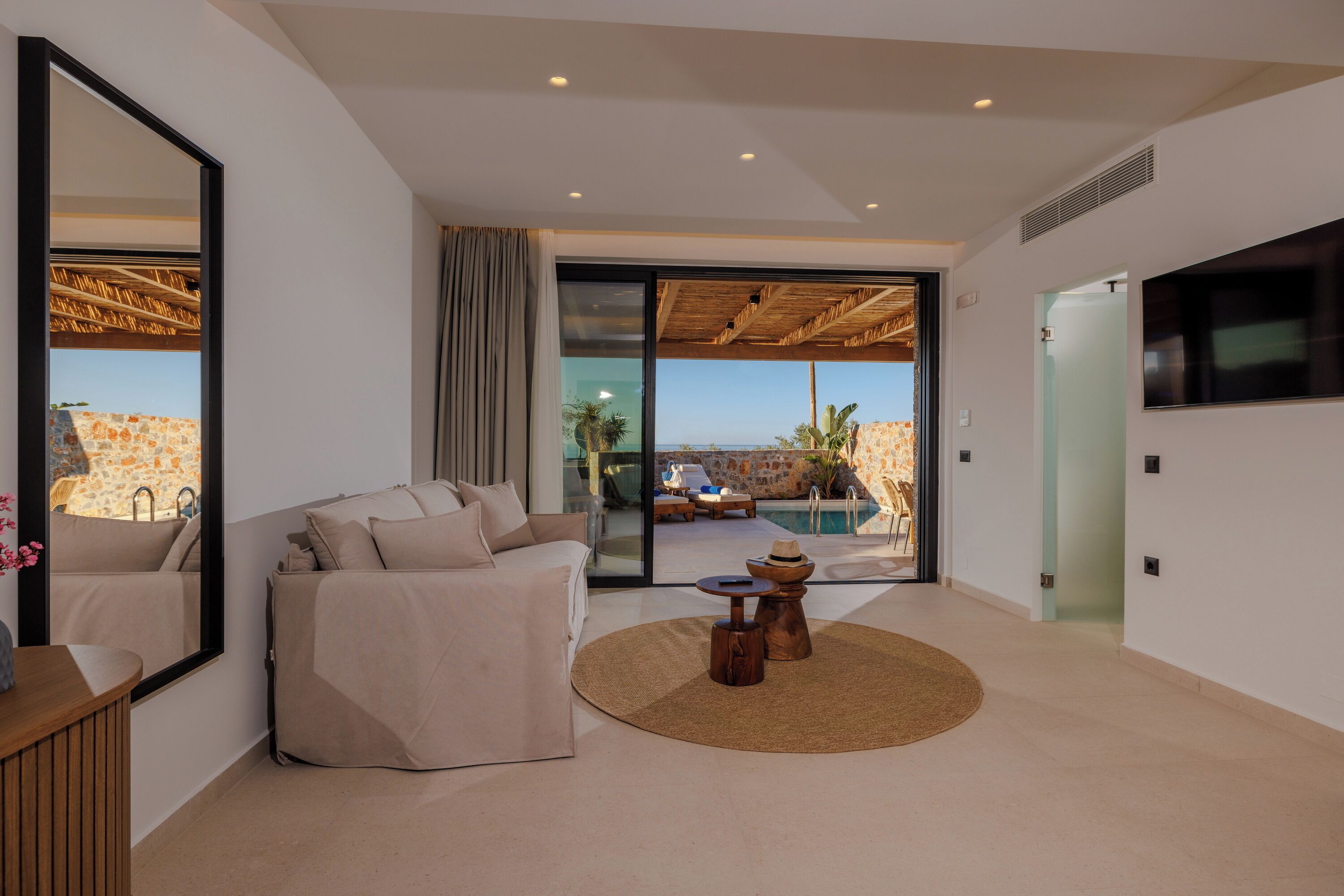 Sala de estar moderna com um espelho grande, sofá bege, mesa de centro redonda e vista para uma piscina e área externa.