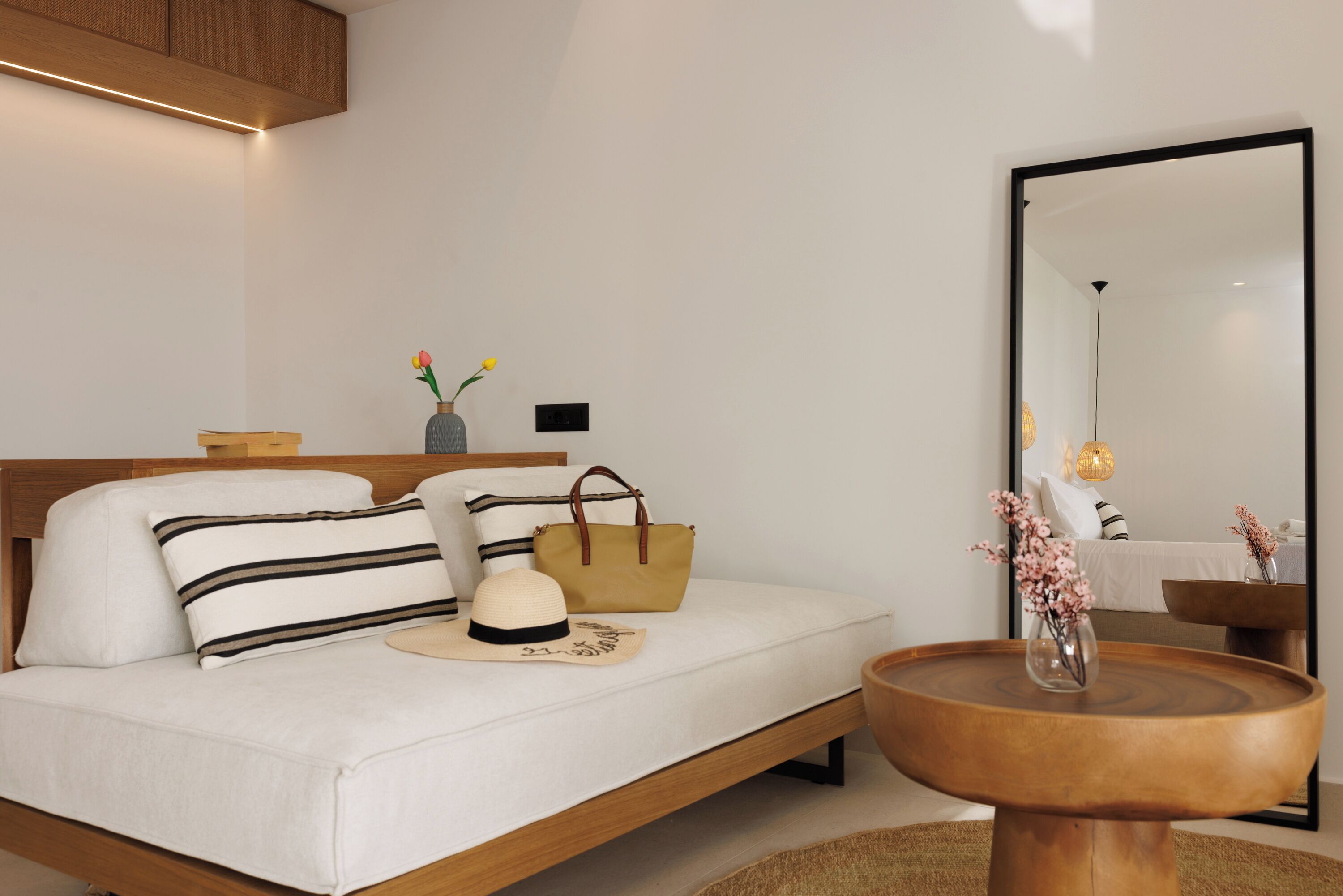 A modern bedroom with a wooden bed frame, white bedding, a bedside table with a vase, and a large mirror.