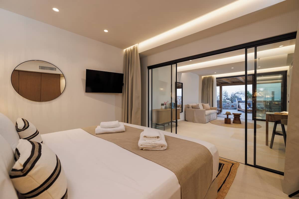 A modern hotel room with a large bed, a flat-screen TV, a sliding glass door leading to a balcony, and a view of the city.