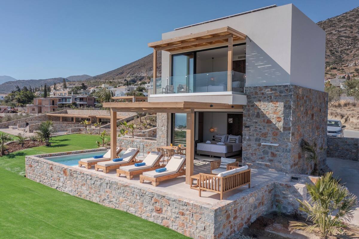 A modern two-story house with a stone exterior, a balcony with lounge chairs, and a swimming pool.