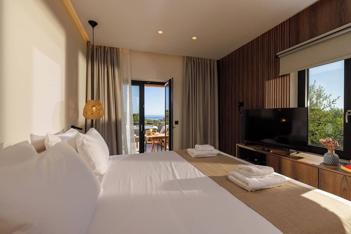 A hotel room with a large bed, a TV, a balcony with a table and chairs, and a view of the outdoors.